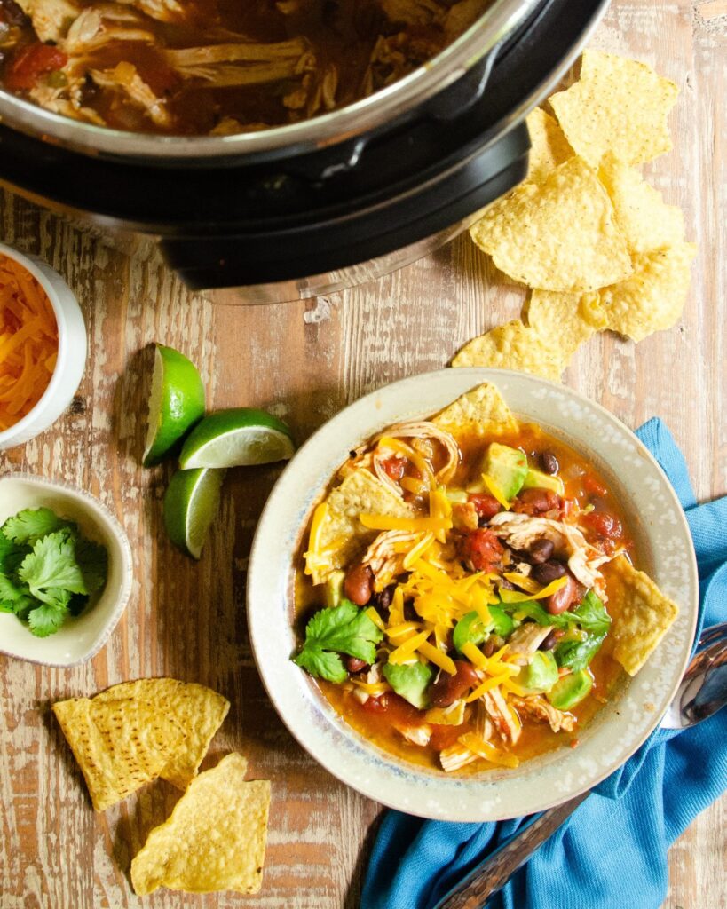A bowl of chicken tortilla soup on a table next to a pressure cooker, along with chips, limes and a blue napkin.