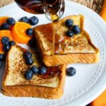 French toast with blueberries and mandarins and maple syrup being poured on top.