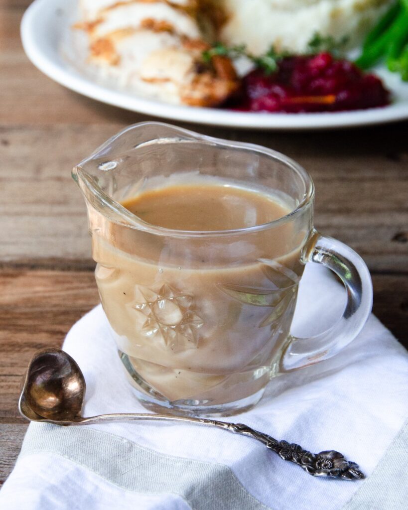 Gravy in a sauce boat on a wooden table with a dinner plate in the background.
