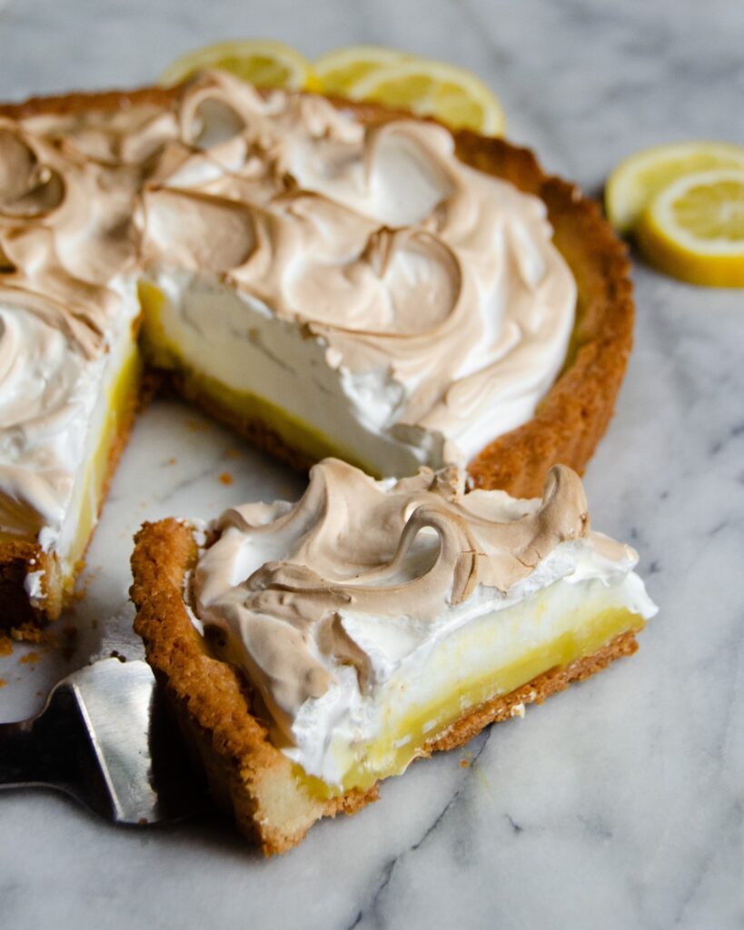 A piece of lemon meringue tart with the rest of the tart in the background on marble.