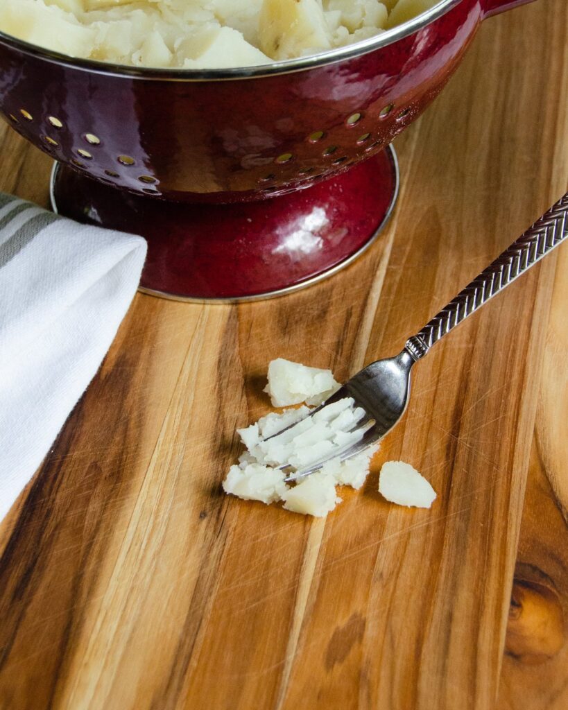 A fork crushing a potato on a board with a colander behind.