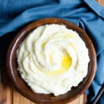 A wooden bowl with mashed potatoes topped with butter and a blue napkin on the side.