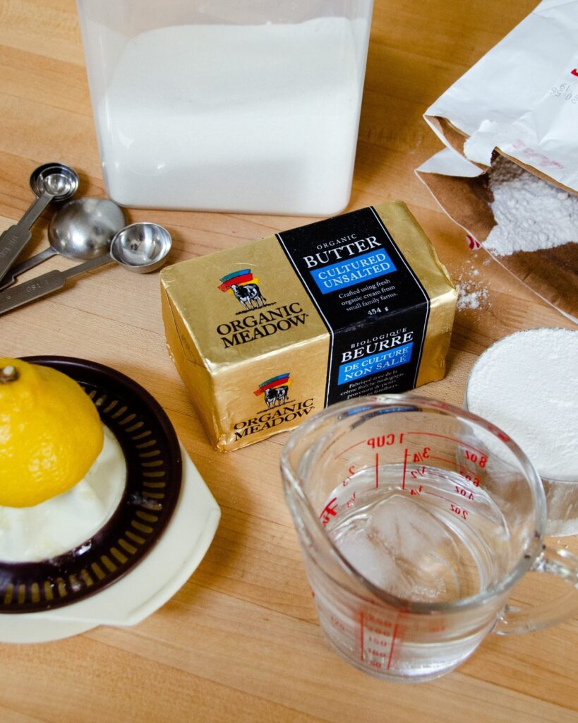 Pastry ingredients on a wooden counter - cultured butter, lemon juice, water, flour, sugar and salt.
