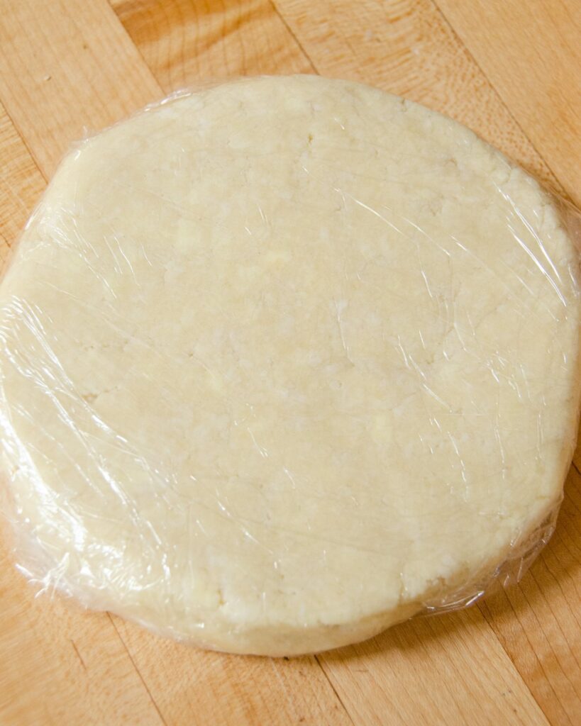 A disk of pastry wrapped in plastic wrap on a wooden countertop.
