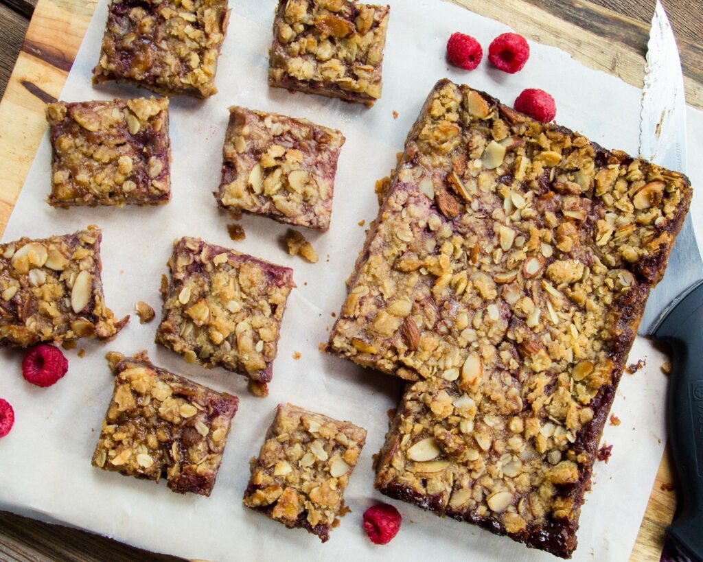 Raspberry Almond Fruit squares cut into pieces on parchment paper on a cutting board with a knife and raspberries around.