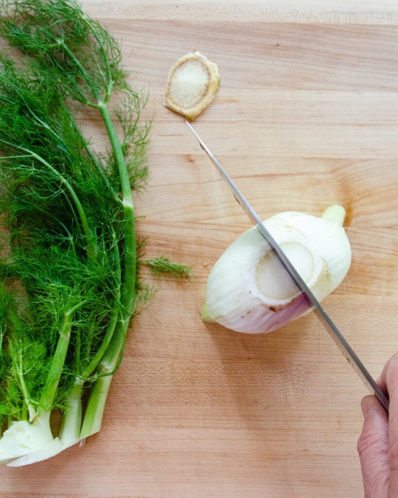 A knife cutting fennel in half on a wooden cutting board.