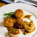 Pan-Seared Scallops with orange tarragon sauce on a white plate with knife and fork in background.