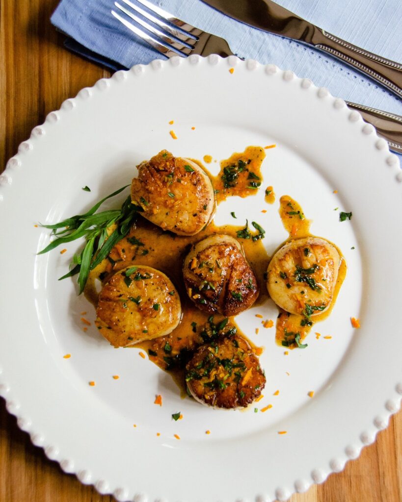 Pan-Seared Scallops on a white plate with orange tarragon sauce drizzled on top.