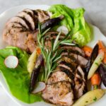 Sous Vide Pork Tenderloin with ginger, honey and rosemary sliced and on a white platter with carrots and rosemary garnish.