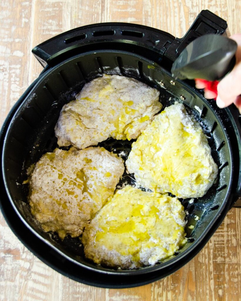 Coated chicken pieces in an air fryer basket being sprayed with oil.