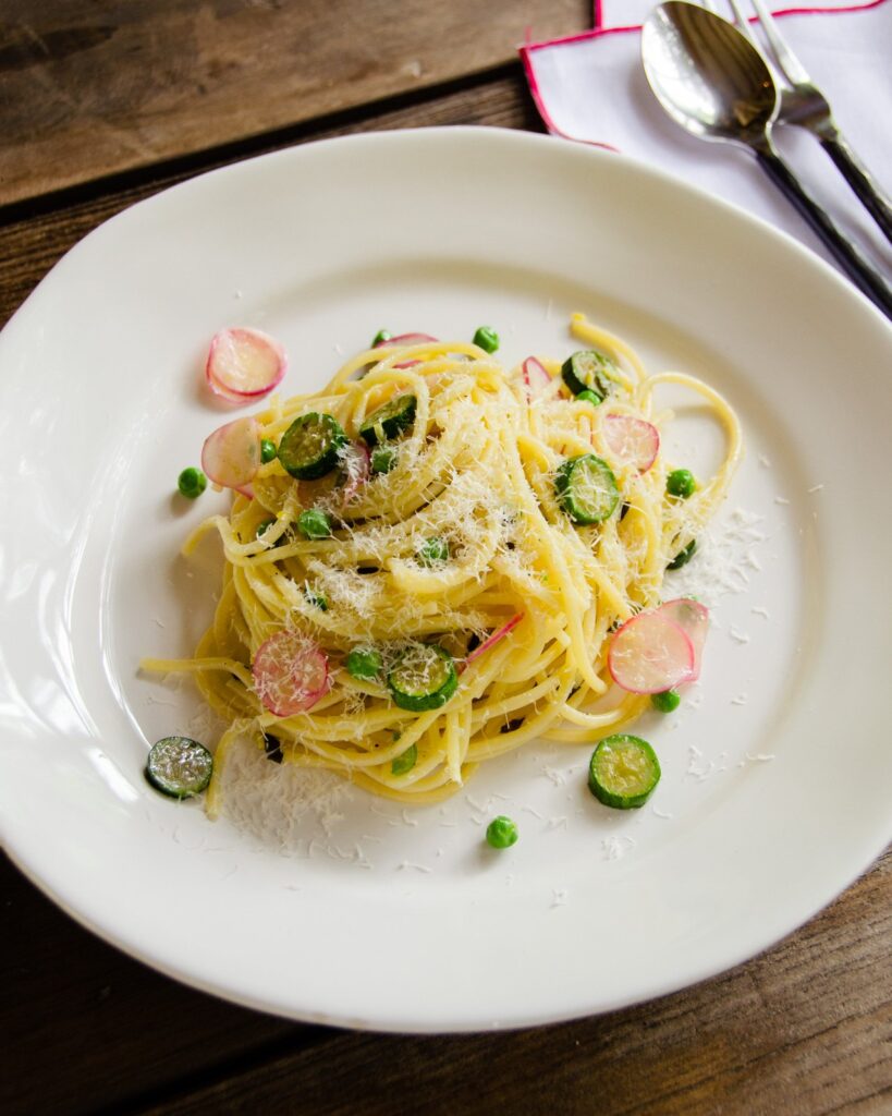 Spaghetti with zucchini, peas, radishes and mint on a white plate on a wooden table with parmesan cheese on the side.