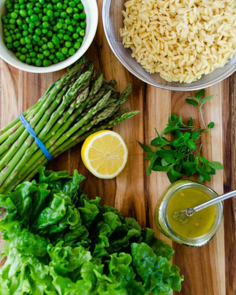 Ingredients for spring green salad on a wooden cutting board - orzo, peas, asparagus, green leaf lettuce, lemon, mint and dressing.