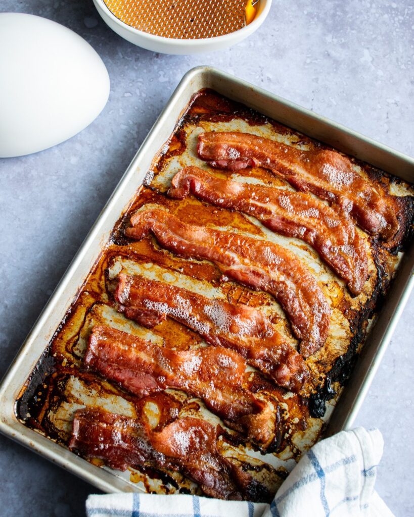 Cooked bacon on a quarter sheet pan with a bacon egg next to it to catch the bacon grease.