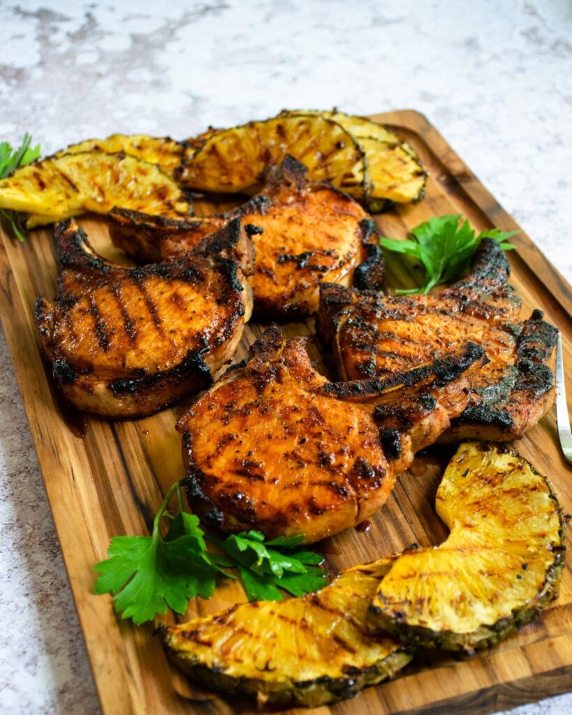 Grilled pork chops on a wooden board with pineapple slices and parsley.