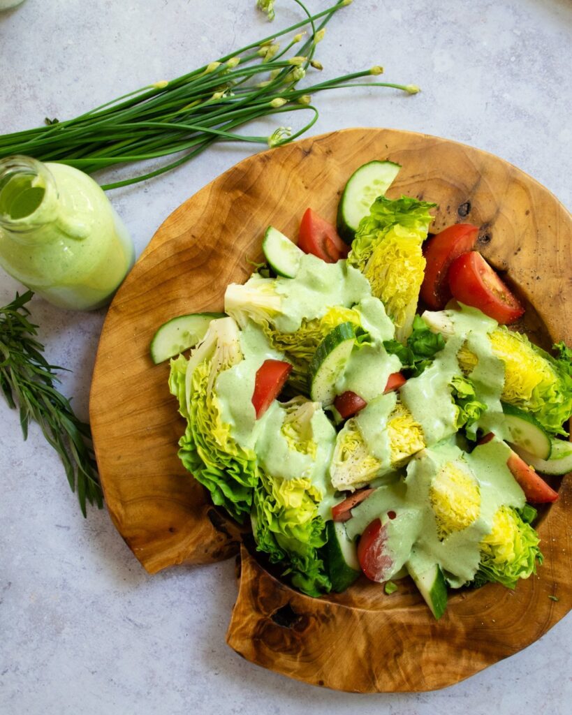 Green Goddess Dressing over baby gem lettuces on a wooden plate with a bottle of dressing and some herbs on the side.