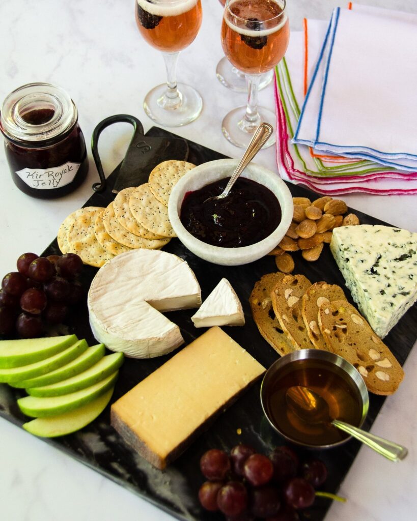 A cheese plate with crackers, apple, grapes and kir royale blackberry jelly with two kir royale cocktails in glasses.