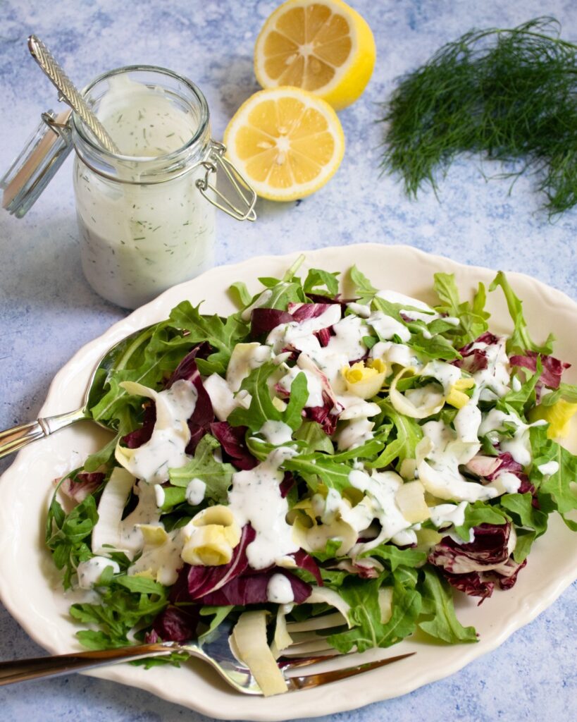 Salad greens dressed on an oval plate with an open jar of lemon dill buttermilk dressing, a halved lemon and some fresh dill on a blue marble counter.