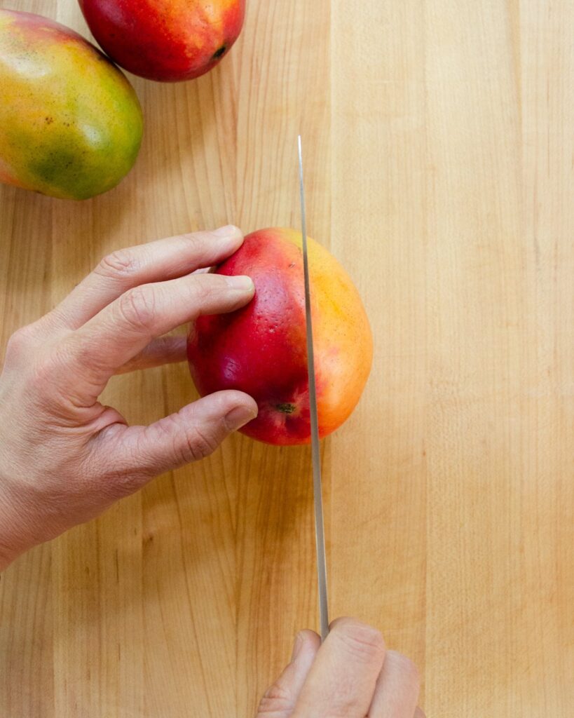 A hand holding a mango and a knife cutting down one side of the mango.