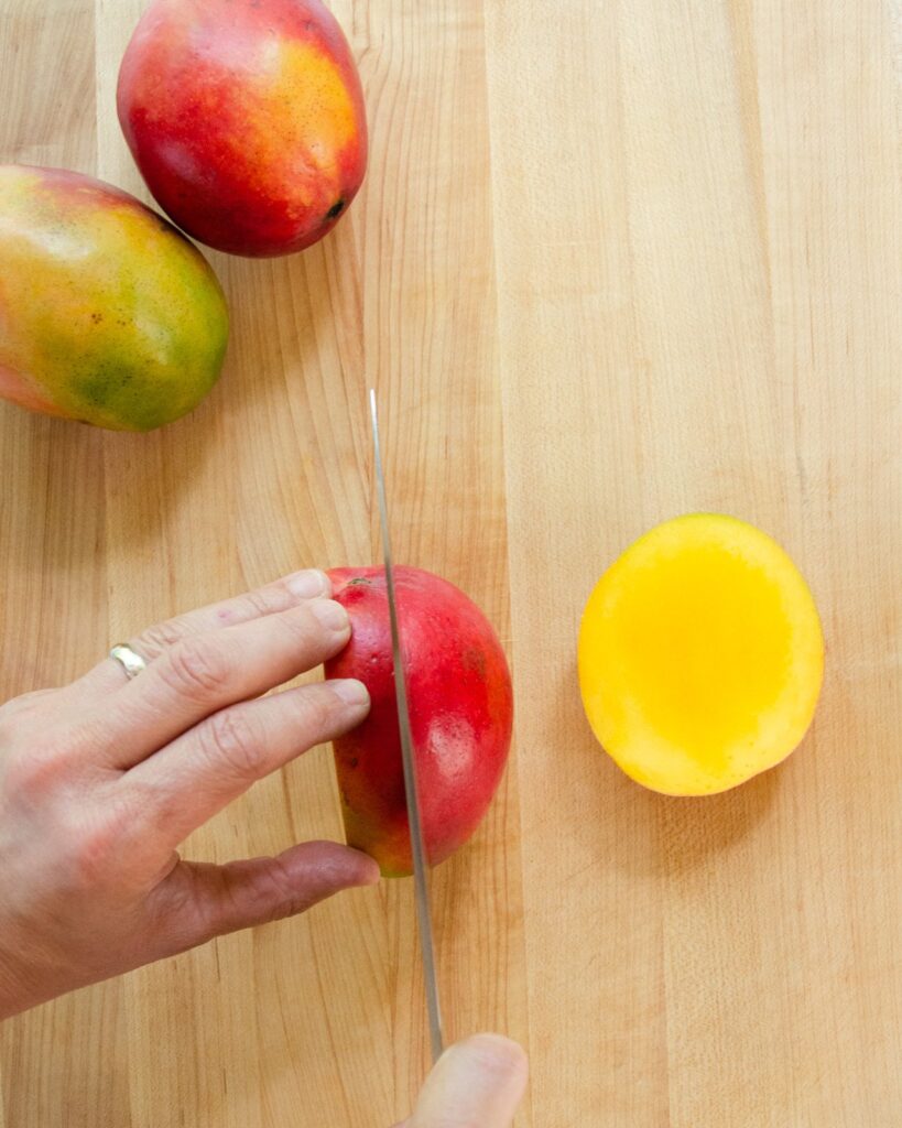 A hand holding a mango and a knife cutting down one side of the mango seed while the other half is sitting on the cutting board.