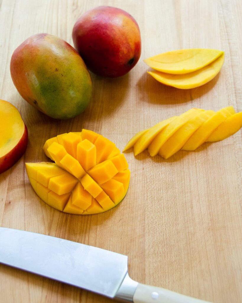 A mango half inverted to show squares of mango next to slices of mango and a chef's knife.