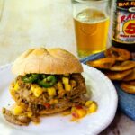 Mango Jalapeno pulled pork sandwich on a white plate with a beer in the background and some chips on the side.
