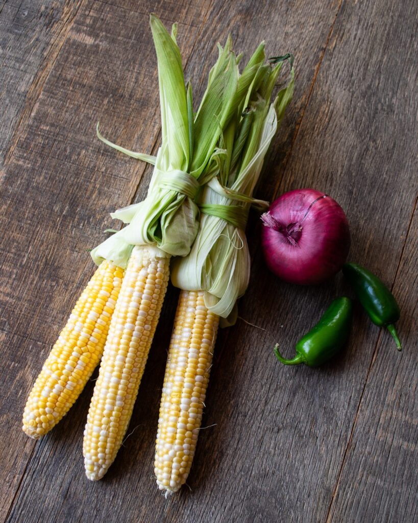 Three ears of corn, a red onion and some Jalapeño peppers on a wooden table.