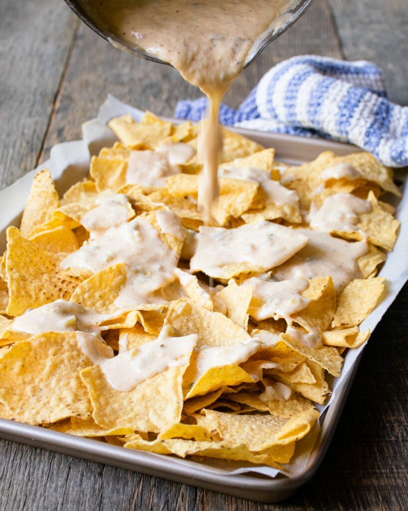 Pouring quest blanco onto corn tortilla chips on a baking sheet.