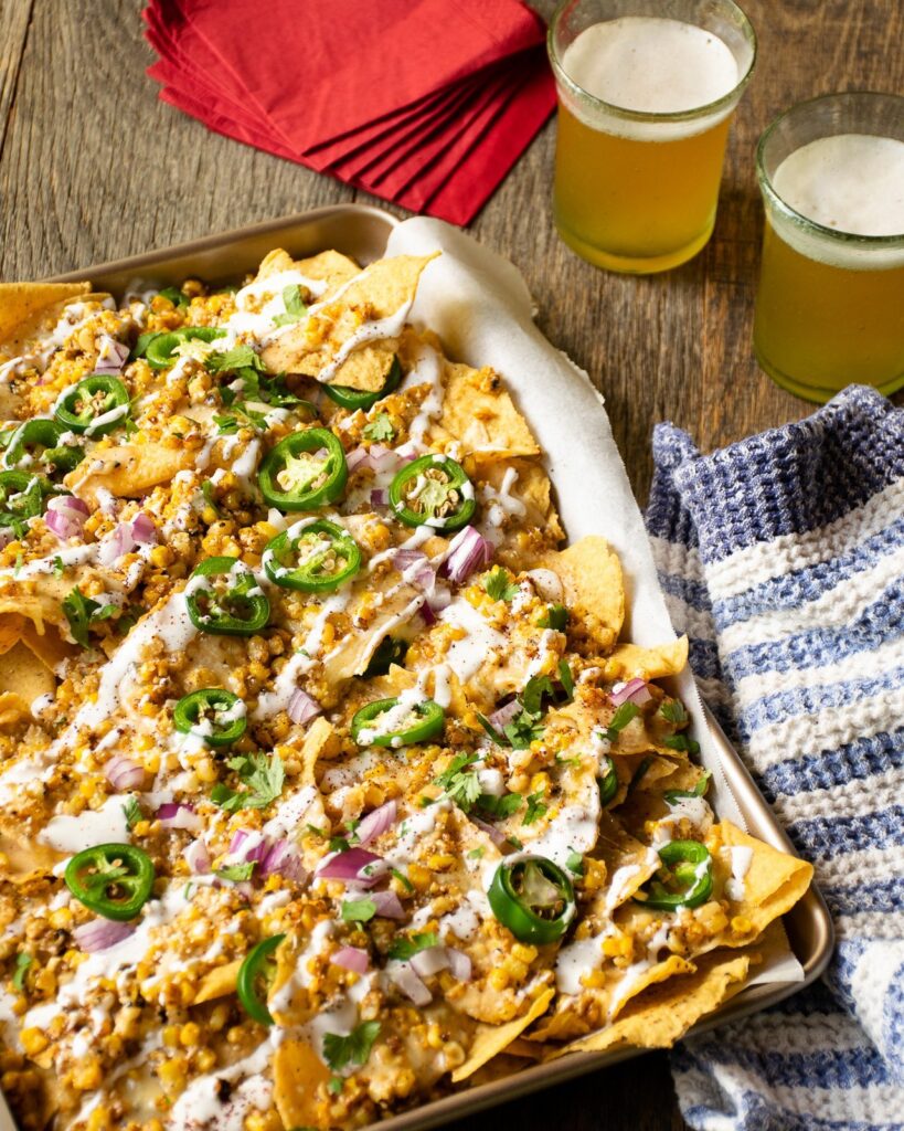 Mexican street corn nachos on a parchment lined sheet pan with two beers and red napkins near by.