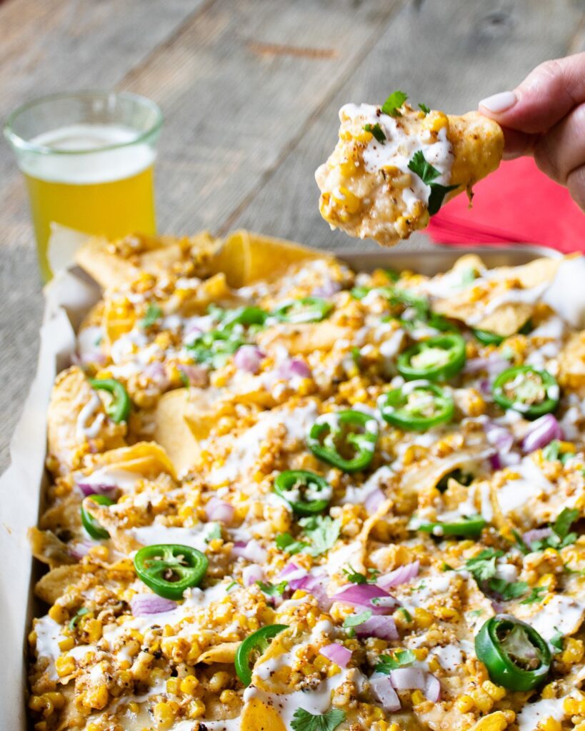 A hand lifting a tortilla chip from a sheet pan of Mexican street corn nachos.