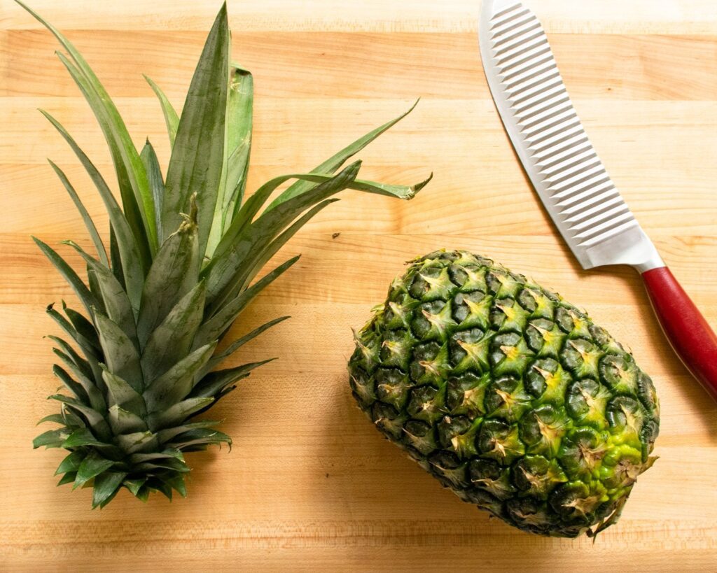 A pineapple with the leaves removed on a cutting board with a red-handled knife.