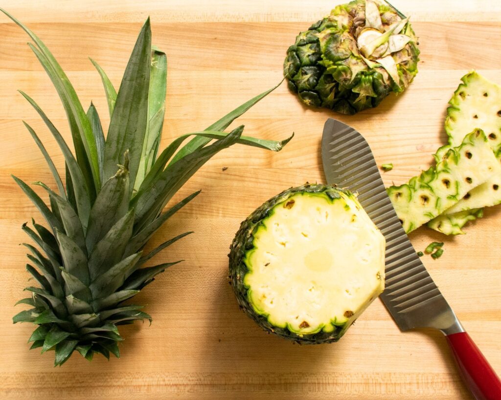 A pineapple with a red-handled knife slicing off the tough outer peel - how to cut a pineapple.