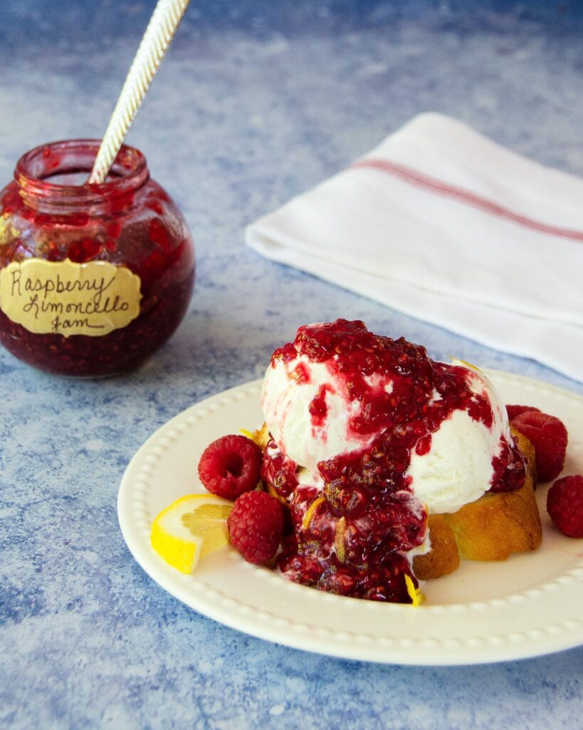 A white plate with pound cake, vanilla ice cream and raspberry limoncello jam on top, with a jar of jam behind.