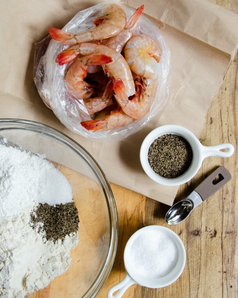 Shrimp on brown paper with  a bowl of cornstarch, salt and pepper on a wooden counter.