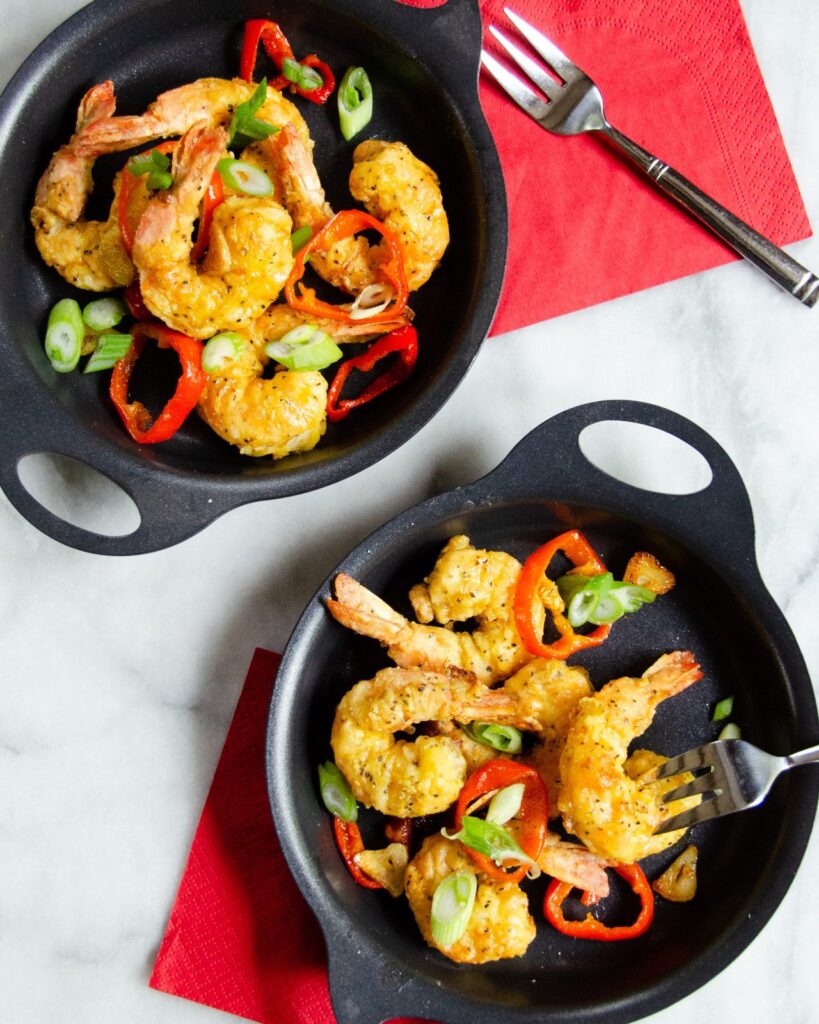 Salt and pepper  shrimp in black dishes with sliced scallions  and cherry peppers on marble with red napkins.