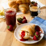 Strawberry jam on a croissant with strawberries scattered around, extra croissants in the background and a cup of espresso.