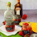 three jars of strawberry-mango margarita jam on a counter with a bottle of tequila and triple sec in the background.