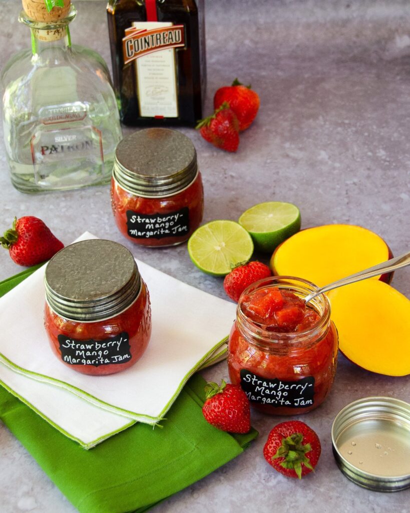 three jars of strawberry mango margarita jam with strawberries, mango, lime and bottles of tequila and triple sec in the background.