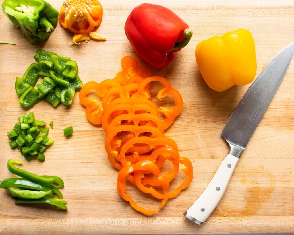 A green pepper cut into pieces and pan orange pepper cut into rings with a white handled chef's knife on a cutting board.