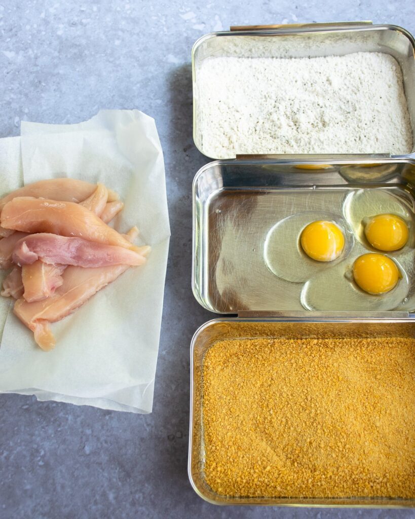 Three dredging pans set up with breadcrumbs, eggs and flour and raw chicken on the side.