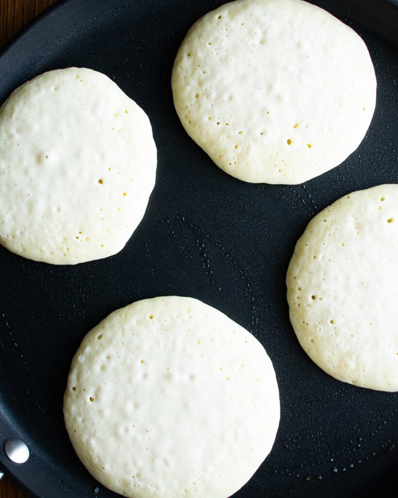 4 pancakes on a griddle with bubbles in the batter read to pop.