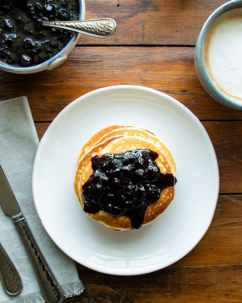 Stack of pancakes with blueberry cinnamon compote on top on a white plate on a wooden table.