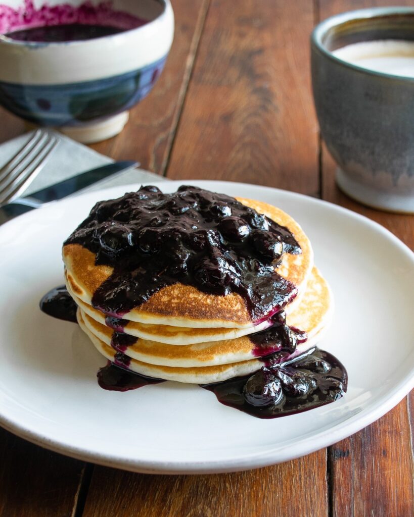 Stack of pancakes with blueberry cinnamon compote on top on a white plate on a wooden table with coffee and a bowl of compote in the background.