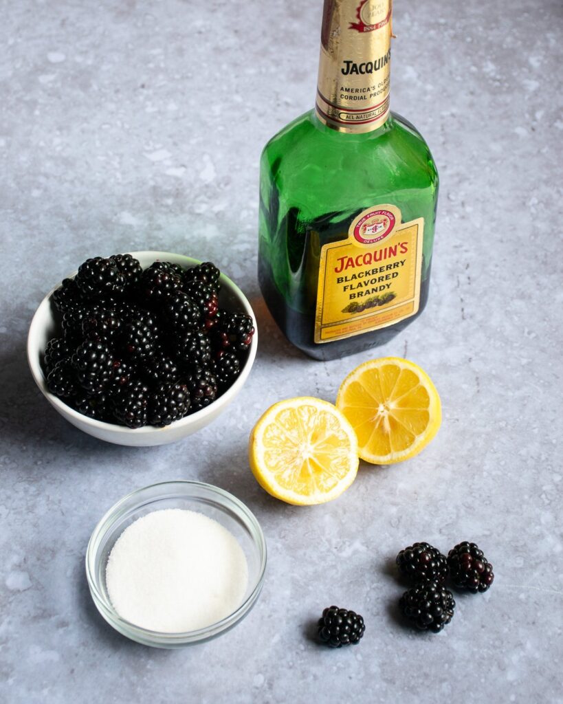 Blackberry Brandy Sauce ingredients - blackberries, lemon, sugar and blackberry brandy - all on a cement counter.