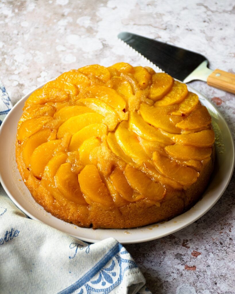 Upside down peach cake on a white platter with a pie server and a blue and tan towel.