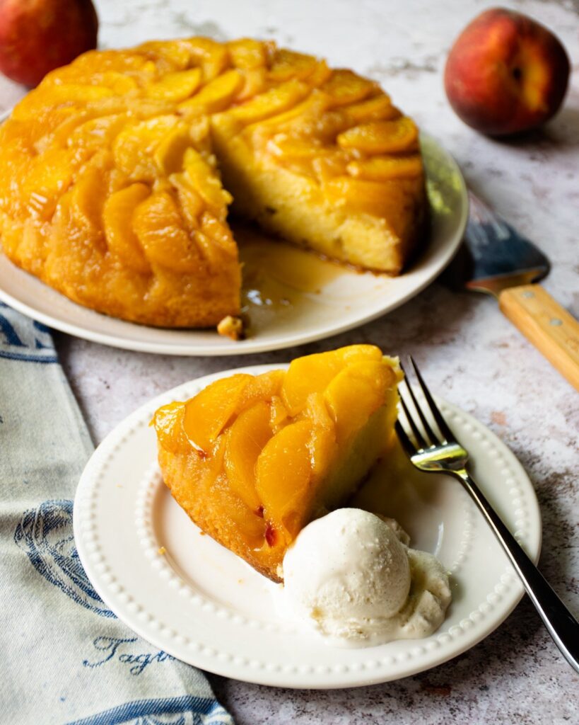 A piece of peach cake on a plate with a scoop of ice cream, with the whole cake behind it.