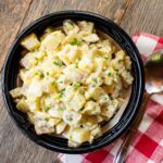 A black ceramic bowl with potato salad on a rustic wooden table with a red and white checkered napkin and spoon.