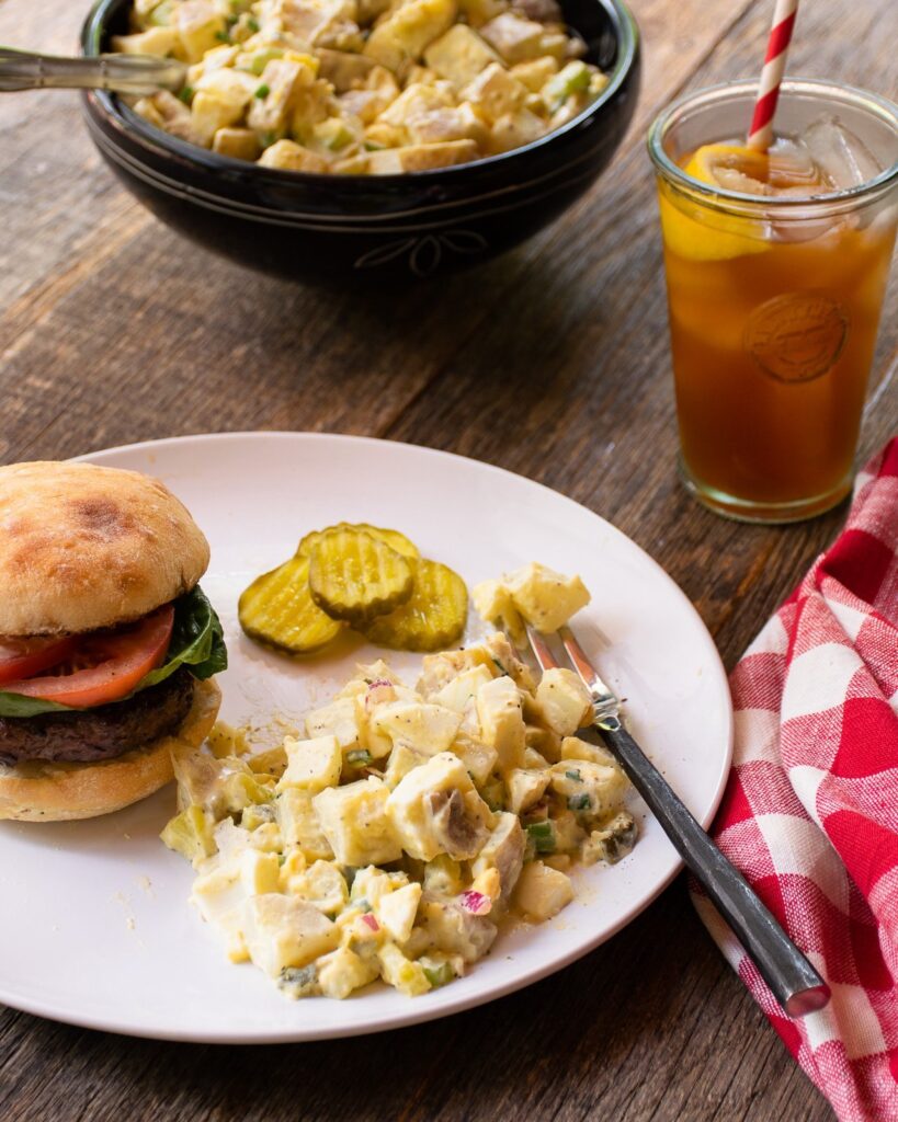 Potato salad with a burger and pickles on a wooden table with more potato salad in the background and a glass of iced tea.