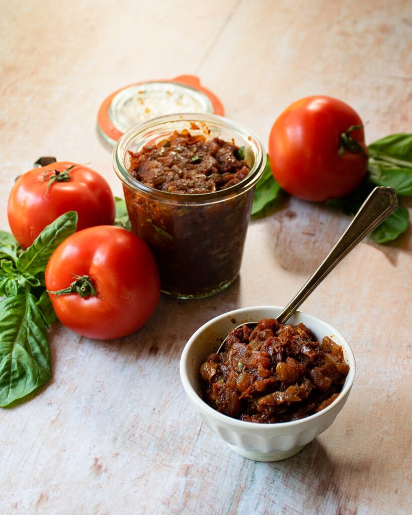 Tomato jam in a little dish with  the open jar and a few tomatoes behind it.