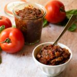 An open jar of tomato jam with a small dish of jam with a spoon on a wooden table with a couple of tomatoes and fresh basil around.