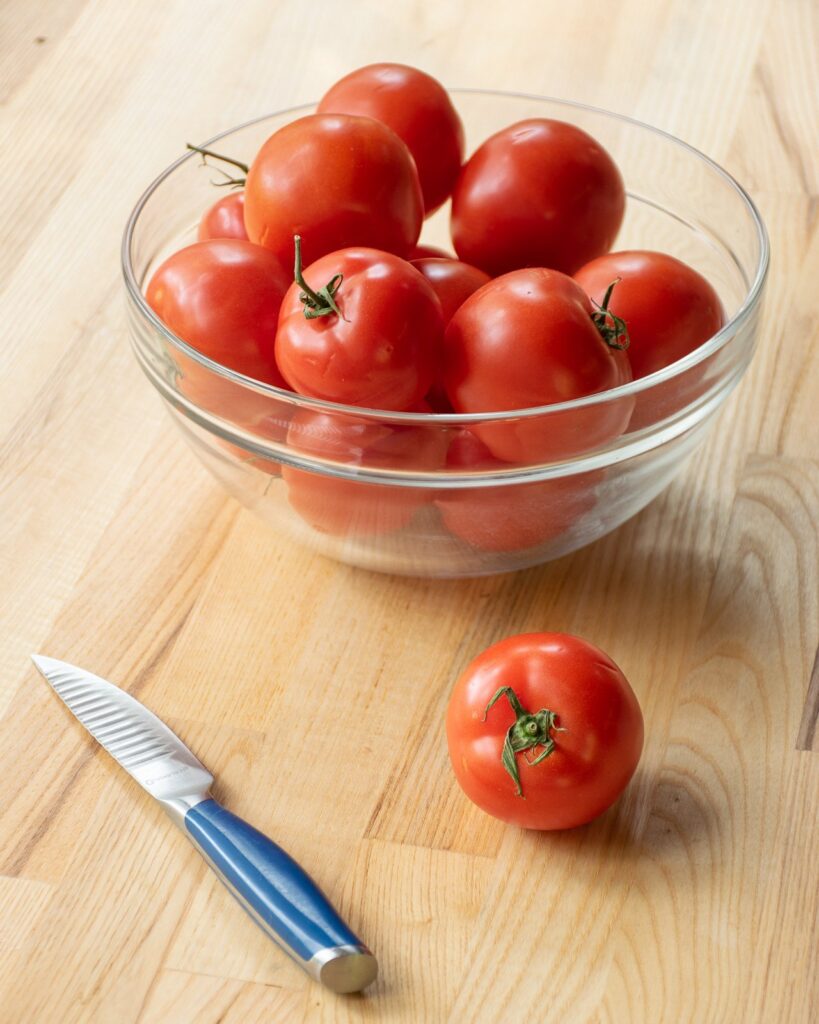 A glass bowl of tomatoes with a single tomato on a cutting board with a paring knife.